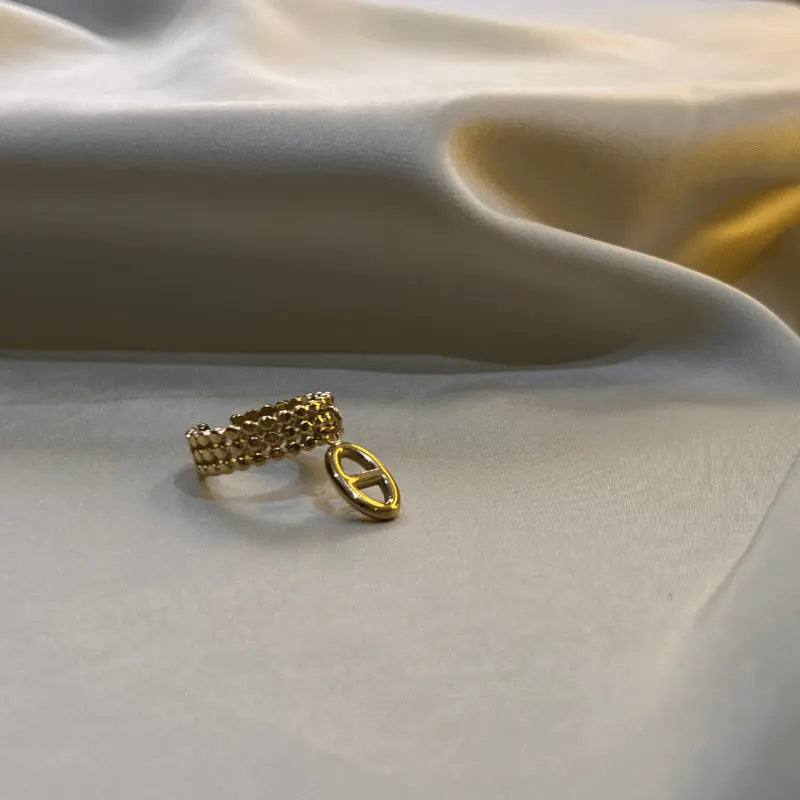 Bague breloque en acier inoxydable doré avec un design en maillons et un pendentif ovale, posée sur un fond en tissu.
