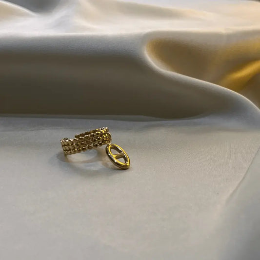 Bague breloque en acier inoxydable doré avec un design en maillons et un pendentif ovale, posée sur un fond en tissu.