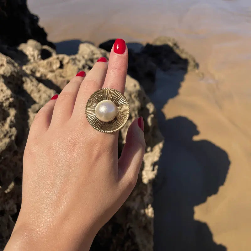 Bague en acier inoxydable dorée avec une perle, portée sur une main avec un arrière-plan de plage