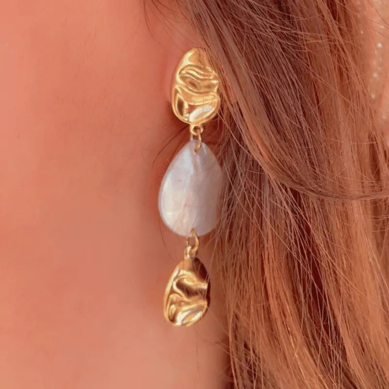 Boucle d'oreille pendante en acier inoxydable dorée avec détail en nacre, portée sur une oreille