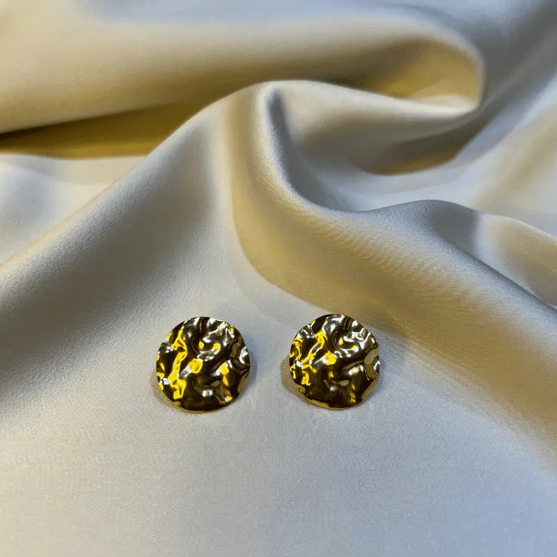 Gros plan de boucles d'oreilles en acier inoxydable texturé, posées sur un fond en tissu satiné