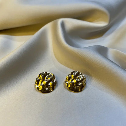 Gros plan de boucles d'oreilles en acier inoxydable texturé, posées sur un fond en tissu satiné