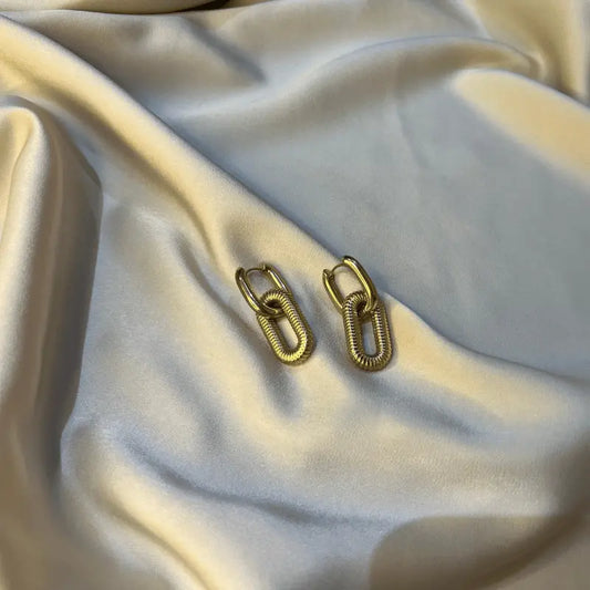 Boucles d’oreilles en acier inoxydable doré avec design de chaînes, posées sur un tissu lisse.