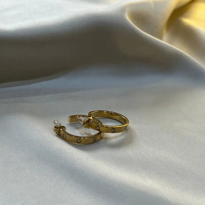 Boucles d'oreilles créoles dorées en acier inoxydable, avec petites pierres brillantes, posées sur un fond en tissu.