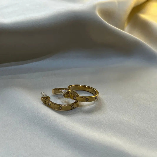 Boucles d'oreilles créoles dorées en acier inoxydable, avec petites pierres brillantes, posées sur un fond en tissu.