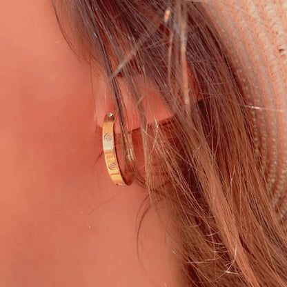 Boucles d'oreilles créoles dorées en acier inoxydable, serties de pierres étincelantes, portées en extérieur.
