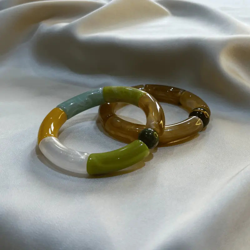 Deux bracelets en acétate, l'un multicolore et l'autre marron, disposés ensemble sur un fond de tissu.