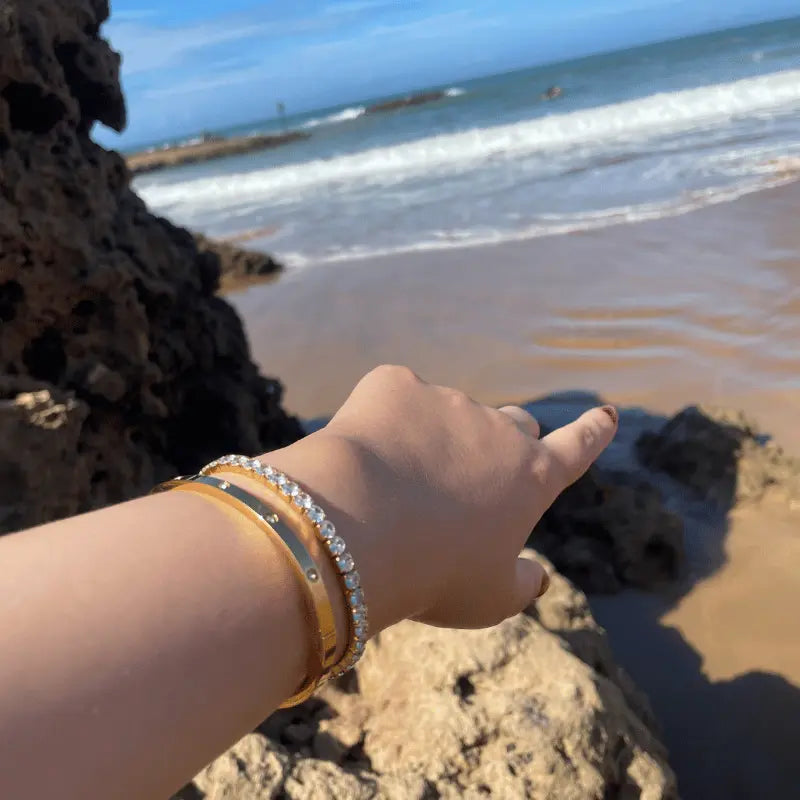 Bracelet rigide en acier inoxydable doré porté avec un bracelet de pierres, en extérieur avec vue sur la plage