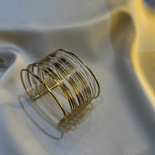 Bracelet manchette large dorée en acier inoxydable avec un design moderne, posée sur un tissu doux pour un look élégant et raffiné.